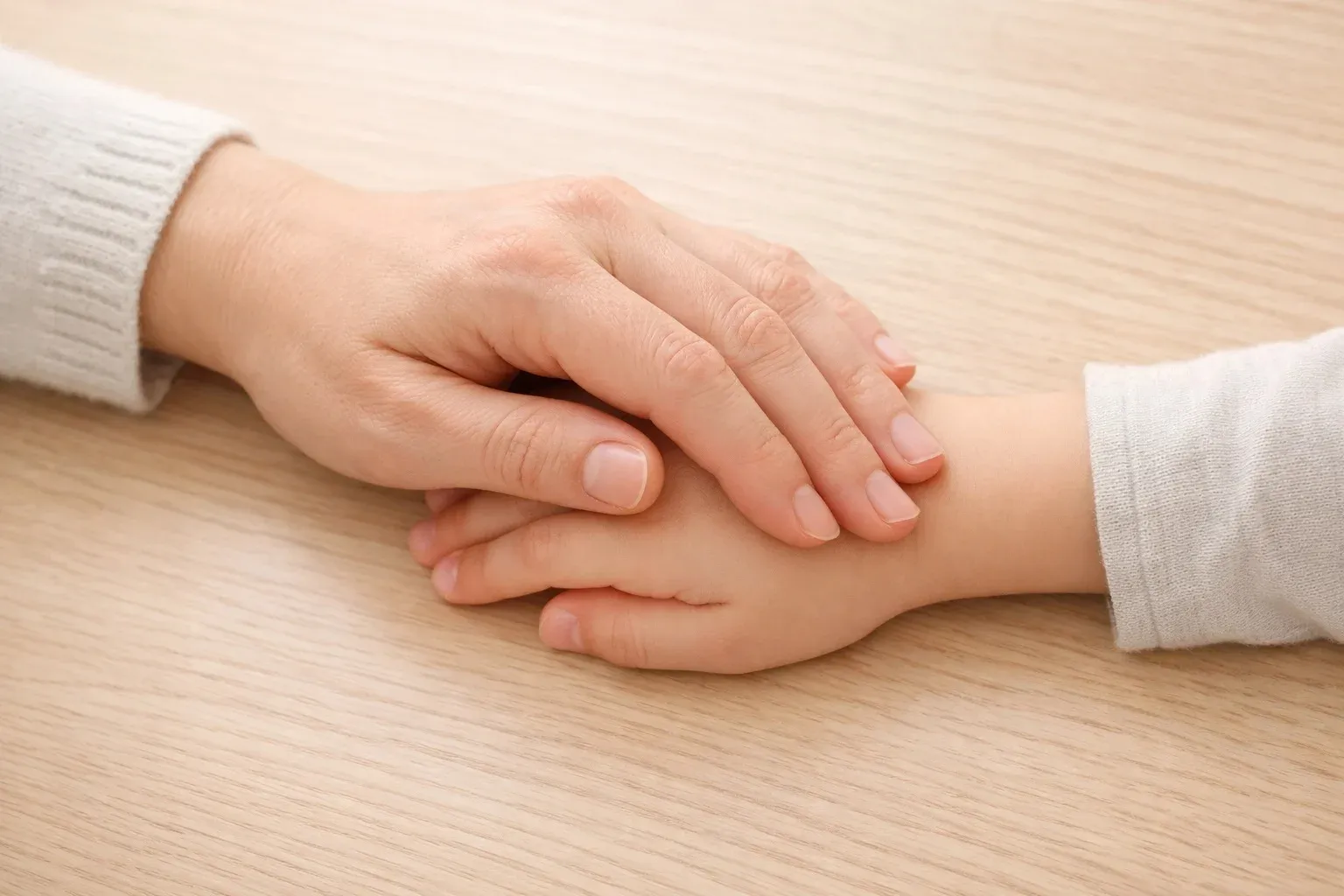 Overhead view of an adult hand gently resting over a child's hand on a wooden table in soft natural light