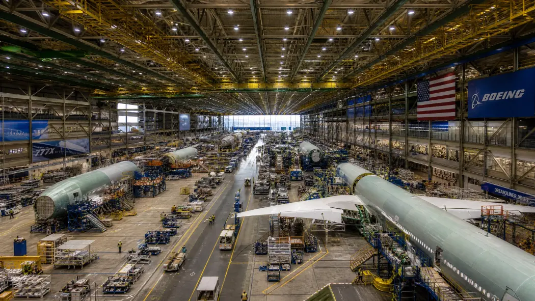 A high-angle interior view of the Boeing Everett Factory assembly line, showing multiple massive aircraft fuselages being built within a gargantuan industrial space.