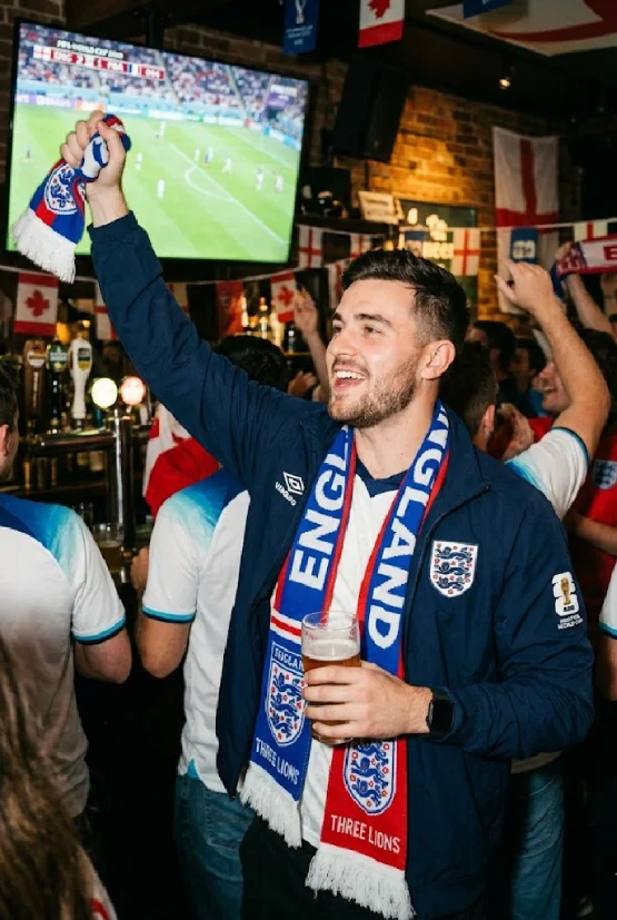 A cheerful man in a navy England national team jacket and scarf celebrating a match victory in a crowded pub during the 2026 FIFA World Cup, holding a beer and a supporter's scarf.